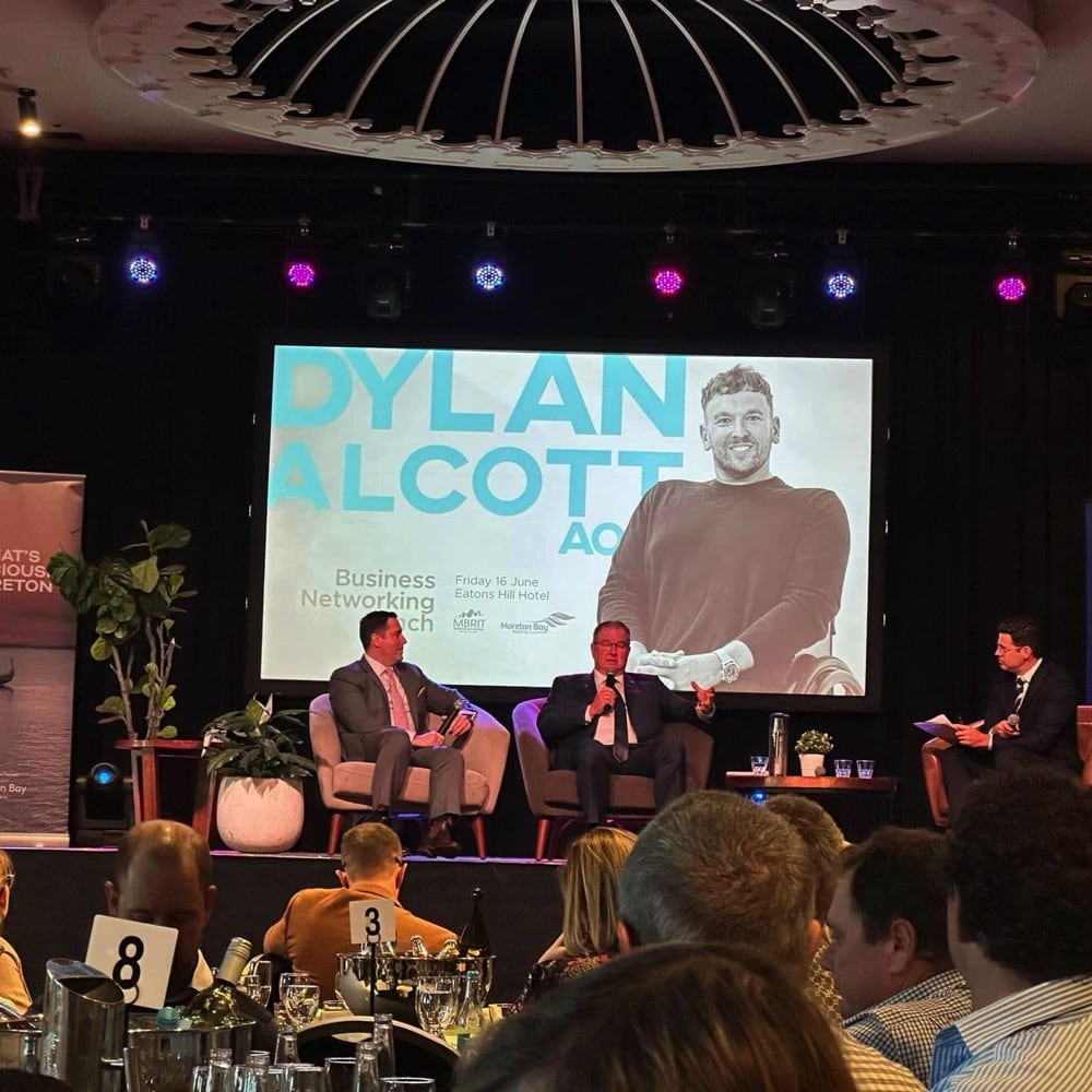 3 men on a stage in lounge chairs talking. A large screen is in the background with a picture of Dylan Alcott on it. People in the foreground are sitting around tables. Live event audio Brisbane, Gold Coast, Sunshine Coast